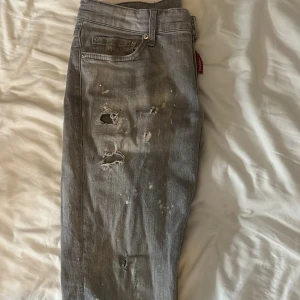 Grå Dsquared2 jeans med slitningar - Säljer ett par grå jeans från Dsquared2 med coola slitningar, hål och färgstänk för en riktigt edgy vibe. Klassisk femficksmodell med rakt snitt och låg midja. Jeansen har en tydlig streetkänsla och är tillverkade i bomull.