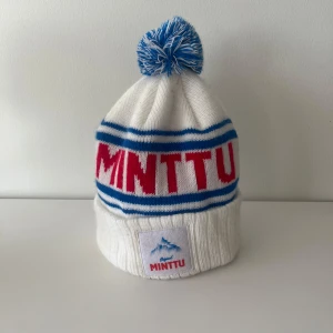 Vit Minttu mössa med blå tofs - Säljer en vit stickad Minttu-mössa med blå och röda detaljer. Mössan har blå ränder, röd text och en stor blå-vit tofs på toppen. Framtill finns en patch med Minttu-loggan och bergmotiv. Perfekt för kalla dagar och snygg att bära på vintern.