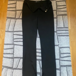 Svarta byxor med slits och knappar - Snygga svarta byxor med unik slits nertill på benen och dekorativa knappar i midjan. Byxorna har en rak passform och är perfekta för dig som gillar stilrena plagg med en twist. Materialet känns mjukt och följsamt.