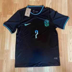 Svart Brasilien fotbollströja Nike - Snygg svart Brasilien fotbollströja från Nike med turkosa detaljer och nummer 7 på bröstet och ryggen. Vini Jr. står tryckt på ryggen. Kortärmad modell med rund hals och turkos kant vid ärmar och nacke.