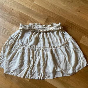 Beige volangkjol med resårmidja - Supersöt beige kortkjol med volanger och bred resår i midjan. Kjolen har en luftig och flowy känsla, perfekt för varma dagar. Den är tillverkad i ett mjukt bomullstyg som känns skönt mot huden och ger en avslappnad vibe.