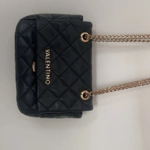 Svart quiltad axelväska från Valentino - Snygg svart axelväska från Valentino med quiltat mönster och guldfärgade kedjor. Väskan har ett lock med magnetlås, innerfack med dragkedja och plats för det viktigaste. Perfekt storlek för att bära med sig över axeln och ger en lyxig känsla till din outfit.