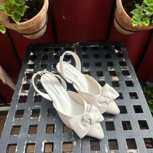 Vita pumps med rosett från Bubbleroom - Snygga pumps i ljusbeige/vit ton från Bubbleroom med spetsig tå och stor rosett framtill. Skorna har en ribbad textur och öppen häl med rem. Perfekta för dig som vill sticka ut med en feminin och trendig look.