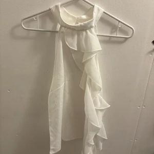 Vit ärmlös volangtopp med halterneck - Säljer en vit ärmlös topp med halterneck och snygga volangdetaljer längs ena sidan. Toppen har en elegant, asymmetrisk design och knäpps i nacken med en liten knapp. Perfekt för dig som gillar stilrena och trendiga plagg.