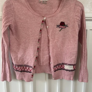 Rosa kofta från Odd Molly med broderi - Superfin rosa kofta från Odd Molly med broderad ros på bröstet och dekorativa detaljer längs knäppningen och nederkanten. Koftan har långärmade ärmar med hålmönster och vita knappar. Perfekt för dig som gillar unika plagg med snygga broderier.