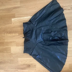 Svart skinnkjol med vid form - Säljer en svart kjol i skinnimitation med snygg, vid och svängd siluett. Kjolen har en hög midja och är kort i modellen, perfekt för att skapa en cool och edgy look. Materialet ger en glansig finish och känns mjukt mot huden.