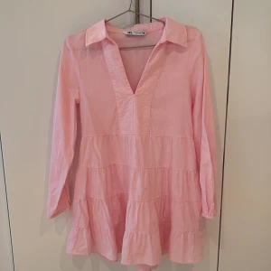 Rosa kortklänning från Zara - Supersöt rosa klänning från Zara med volanger och v-ringad hals. Klänningen har lång ärm och krage, samt är tillverkad i mjuk bomull. Perfekt för dig som gillar en feminin och chill stil.