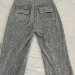 Grå Juicy Couture mjukisbyxor S - Grå mjukisbyxor från Juicy Couture med bred resår och snörning i midjan. Byxorna har två fickor fram och två bakfickor med lock och knappar. Mjukt material och diskret logga broderad på ena fickan. Perfekta för chill dagar.