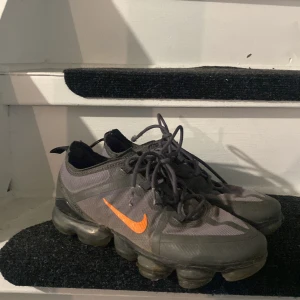 Nike Vapormax sneakers grå/orange - Nike Vapormax sneakers i mörkgrått med orange swoosh på sidorna. Skorna har en unik bubblig sula, snörning och är tillverkade i syntetmaterial och mesh för en sportig vibe. Perfekta för dig som gillar streetstyle och vill sticka ut med coola detaljer dom är i helt okej skick. 