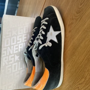Golden Goose sneakers svart/orange - Snygga Golden Goose sneakers i svart mocka med vit stjärndetalj på sidan och orange läderdetalj vid hälen. Klassisk rund tå, vita snören och platt sula. Perfekta för dig som gillar streetstyle med en unik touch. Kommer med orginal box