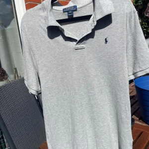 Grå pikétröja från Polo Ralph Lauren XL 18/20 år - Klassisk grå pikétröja från Polo Ralph Lauren i storlek XL. Tröjan har korta ärmar, krage och knäppning framtill med två knappar. Broderad logga på bröstet. Perfekt basic till garderoben och enkel att matcha.