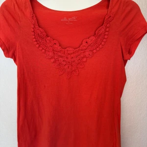 Korallröd topp med virkad spetsdetalj Willi Smith - Snygg korallröd topp från Willi Smith med korta ärmar och v-ringning dekorerad med virkad spetsdetalj framtill. Mjuk och skön bomullskänsla, perfekt till jeans eller kjol när du vill ha något extra. Färgen är korallröd och modellen är normal i passformen.