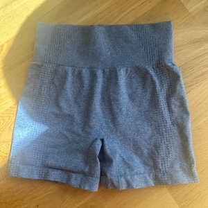 Grå högmidjade träningstights shorts - Snygga grå shorts med hög midja, perfekta för gymmet eller löpning. Stretchigt material med 54% polyamid, 34% polyester och 12% elastan ger en tight och bekväm passform. Diskret mönstrad struktur längs sidorna för extra stil.