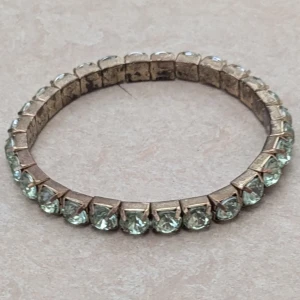 Guldigt armband med ljusa stenar - Snyggt armband i guldton med glittriga ljusa stenar runt hela bandet. Stenarna är infattade i fyrkantiga fästen och ger ett coolt och lyxigt intryck. Perfekt för dig som gillar bling och vill sticka ut med något unikt på handleden.