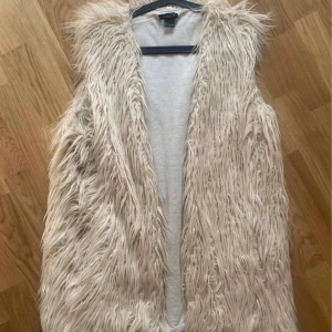 Beige långhårig pälsväst från Lindex - Säljer en supermjuk och fluffig beige pälsväst från Lindex. Västen har långhårig fuskpäls och är öppen framtill utan knäppning. Perfekt för att ge din outfit en cool och trendig touch. Passar dig som gillar statement-plagg och vill sticka ut.