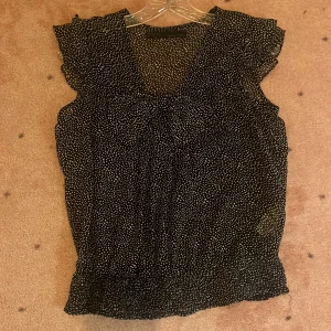 Svart prickig blus med volangärm - Snygg svart/mörkblå blus med små vita prickar och volangdetaljer vid ärmarna. Blusen är ärmlös och har en lätt och luftig känsla, perfekt för varmare dagar. Den har en lös passform och är tillverkad i ett tunt, transparent material. Medium men sitter som S.