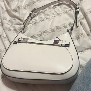 Vit axelväska från H&M i skinnimitation - Säljer en stilren vit axelväska från H&M med snygga silverdetaljer och justerbart axelband. Väskan har en minimalistisk design med mjuka former och magnetlås. Perfekt för dig som gillar clean och modern look.