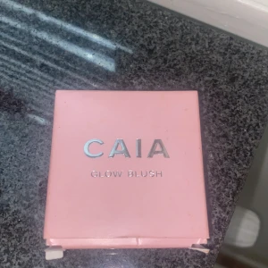 CAIA Glow Blush i persikorosa - Säljer en CAIA Glow Blush i en snygg persikorosa nyans. Kommer i en stilren ljusrosa dosa med spegel i locket. Rouget har en skimrande finish och är pressat i ett cirkulärt mönster. Perfekt för att ge kinderna ett fräscht glow.