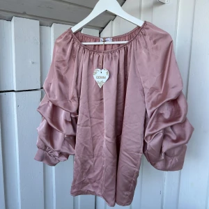 Rosa blus med volangärmar från Gemini - Supersöt rosa blus från Gemini i glansigt material med bred resår i halsen och stora volangärmar. Perfekt för dig som gillar statement-ärmar och en romantisk vibe. Blusen har en lös passform och är lätt att styla till både jeans och kjol.