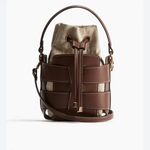 Brun och beige bucket bag med snörning - Snygg bucket bag i brun skinn och beige canvas med cool snörning upptill. Väskan har både handtag och axelrem, perfekt för att bära på olika sätt. Stilren design med kontrastsömmar och detaljer i guld. Rymmer det viktigaste och ger en trendig touch till din outfit.