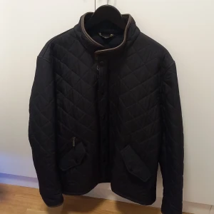 Svart jacka från Barbour - Quiltad svart jacka från Barbour med snygga bruna detaljer vid kragen. Klassisk design med knappar framtill, två fickor med lock och Barbour-broderi. Perfekt för dig som gillar stilren och tidlös look. Perfekt för hösten. Använd ett fåtal gånger i mycket fint skick.