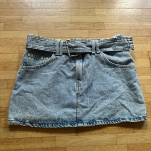 Blå jeanskjol med bälte - Snygg ljusblå jeanskjol från New Yorker med klassisk femficksdesign och tillhörande jeansbälte med metallspänne. Kjolen har en kort och midwaist passform med slitna detaljer vid kanterna för en cool vibe. Perfekt för dig som gillar denim och vill ha en enkel men trendig look.