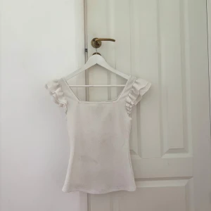 Vit topp med volangärmar - Supersöt vit topp med fyrkantig ringning och volangdetaljer på axlarna. Toppens material känns mjukt och stretchigt, perfekt för en fräsch och stilren look. Passar lika bra till jeans som kjol och ger en romantisk vibe.