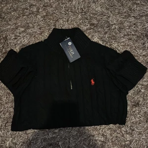 Svart stickad polotröja Polo Ralph Lauren - Svart stickad polotröja från Polo Ralph Lauren med klassisk kabelstickning och röd broderad logga på bröstet. Tröjan har half zip-dragkedja och hög ribbad krage som ger en chill och snygg look. Perfekt för dig som gillar stilrena och tidlösa plagg.