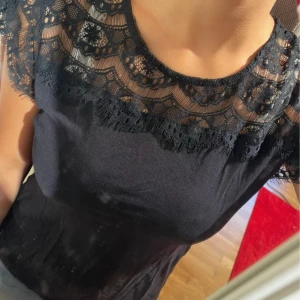 Svart spetsblus från Lindex - Supersnygg svart blus från Lindex med transparent spets upptill och på ärmarna. Blusen har rund halsringning och korta ärmar med vacker spetsdetalj som ger en elegant vibe. Perfekt att styla med jeans eller kjol för en trendig look.