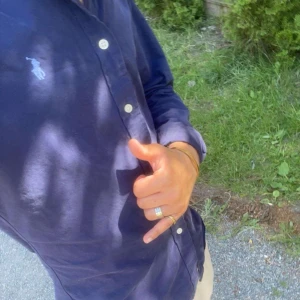 Mörkblå skjorta från Ralph Lauren - Snygg mörkblå skjorta från Ralph Lauren med klassisk passform och vita knappar framtill. Skjortan har lång ärm och den ikoniska polospelaren broderad på bröstet. Perfekt för dig som gillar stilrena och tidlösa plagg.