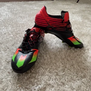 Adidas Messi 15.1 fotbollsskor - Säljer ett par Adidas Messi 15.1 fotbollsskor med snygg design i svart, rött och grönt. Skorna har snörning, mönstrad ovandel och gröna dobbar för grymt grepp på planen. Perfekta för dig som vill sticka ut och spela som Messi. Elite skor 