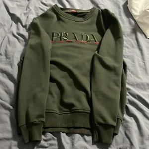 Mörkgrön sweatshirt från Prada - Snygg mörkgrön sweatshirt från Prada med broderad logga i grönt och rött framtill. Tröjan har rund hals, ribbade muddar och är tillverkad i mjuk bomull. Perfekt för dig som gillar exklusiva märken och stilren design.
