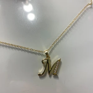 Guldigt halsband med M-berlock - Snyggt halsband från Evry Jeweles i guld med en berlock formad som bokstaven M. Berlocken har glittriga stenar på ena sidan som ger extra bling. Kedjan är tunn och har ett hjärtformat hänge vid låset. Perfekt för dig som vill ha något personligt och stilrent. Nytt kostar 300kr🩷