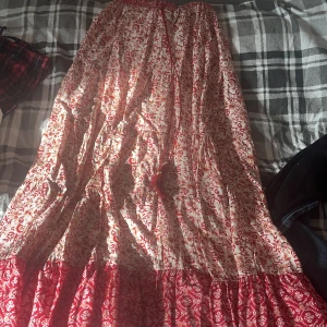 Rödvit långkjol med blommigt mönster - Säljer en flowy långkjol i vitt och rött med blommigt mönster och volangkant nertill. Kjolen har en snygg knytning i midjan med tofsar och är perfekt för dig som gillar boho-stil. Lätt och luftig, passar grymt till sneakers eller sandaler.