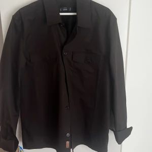 Svart skjorta från MNG Essentials - Stilren svart skjorta från MNG Essentials med klassisk krage och knäppning framtill. Skjortan har två bröstfickor med lock och knappar samt långa ärmar. Perfekt för dig som gillar en clean och enkel look. Nypris 500 säljer för 150