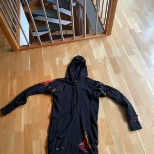 Svart Ed Hardy hoodie med tryck - Svart lång hoodie från Ed Hardy med färgstarka tryck i rött, gult och lila. Framsidan har ett brinnande hjärta, baksidan har stort dödskallemotiv och text. Guldig dödskalle-dragkedja, huva och långa ärmar. Materialet är bomull och modellen är loose fit.