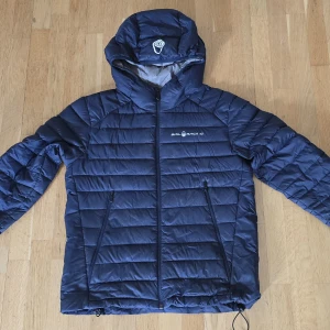 Blå dunjacka från Sail Racing - Säljer en blå dunjacka från Sail Racing, spray down jacket med huva och dragkedja framtill. Strl 170. Jackan är quiltad och har två fickor med dragkedja. Tillverkad i nylon och fylld med certifierat ankdun och fjäder. Perfekt för kalla dagar när du vill hålla värmen och ändå se snygg ut.
