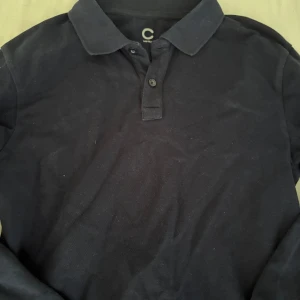 Svart långärmad pikétröja från Cubus - Svart pikétröja från Cubus med klassisk krage och två knappar framtill. Tröjan är långärmad och har en stilren, enkel design som funkar till många olika looks. Materialet känns som bomull och är mjukt mot huden.