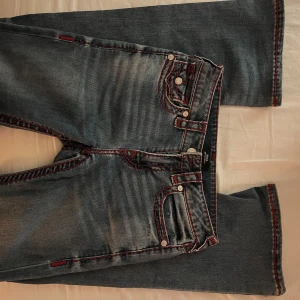 True religon med burgundy färgade sömmar - Säljer ett par blå jeansbyxor från True Religion med tydliga röda kontrastsömmar och klassiska fickdetaljer. Byxorna har bootcut ben och silverfärgade knappar. Perfekta för dig som vill sticka ut med snygga detaljer. Endast använda fåtalet ggr och den enda ”defekten” är lite slit på häl sömmarna som syns på bild. Köpta i USA i november för 2500kr. Midrise i modellen ”Becca”