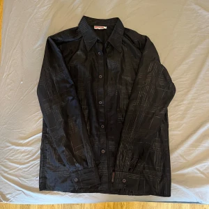 Vintage/äldre Prada skjorta - Säljer en svart skjorta från Prada med diskret rutigt mönster i grått. Skjortan har klassisk krage, knäppning framtill och långa ärmar. Tillverkad i Italien och har en snygg logotypdetalj vid knappraden. Perfekt för dig som gillar stilrena designer. (Inte 100% säker på äktheten då den är köpt för ett par år sedan, därav priset. Men ser väldigt bra ut allmänt så jag skulle ana att den är äkta.)
