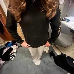 Svart SOL GF hoodie från Clique - Svart hoodie från Clique med huva och magficka. På ryggen finns ett stort guldtryck med sol och texten 'SOL GF', och på ena ärmen står 'GYMNAST' i guld. Perfekt för dig som gillar gymnastik och vill sticka ut med coolt tryck.