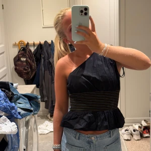 Svart oneshoulder topp med volang - Säljer en svart oneshoulder topp med bred resår i midjan med tunna guldiga ränder. Toppen har volang nertill och en cool asymmetrisk axel. Köpt på zara för 299 kr men oanvänd, storlek S. 