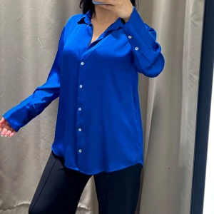 Kornblå skjorta i silkeslook - Säljer en kornblå skjorta med snygg silkesfinish och klassisk krage. Skjortan har långa ärmar, vita knappar framtill och en loose passform som ger en chill vibe. Perfekt att styla med jeans eller dressade byxor för en cool look.