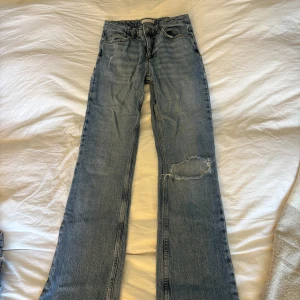 Blåa jeans från Gina tricot, storlek 34 - Snygga ljusblå jeans från Gina tricot i klassisk femficksmodell. Jeansen har raka ben, hög midja och en cool sliten detalj med hål på ena benet. Tillverkade i jeansmaterial med tydliga sömmar och läderpatch bak i midjan.