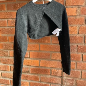 Svart croppad kofta från Gina Tricot - Snygg svart croppad kofta från Gina Tricot med lång ärm och knapp framtill. Koftan har en omlott-design och är stickad i mjukt material som passar perfekt till lager-på-lager. Enkel att matcha med både jeans och kjol för en cool look.