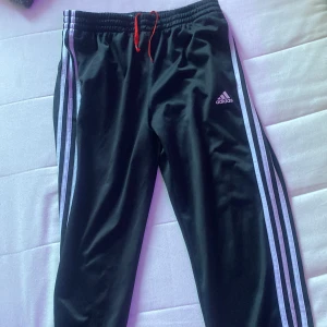 Svarta Adidas mjukisbyxor med ränder - Svarta mjukisbyxor från Adidas med klassiska vita ränder längs sidorna och Adidas-logga på låret. Byxorna har elastisk midja med röd dragsko och är gjorda i ett glansigt syntetmaterial. Perfekta för chill eller träning.