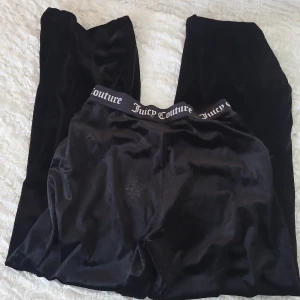 Svarta velourbyxor Juicy Couture - Mysiga svarta velourbyxor från Juicy Couture med bred resår i midjan där loggan är tryckt runtom. Byxorna har en klassisk loose passform och är perfekta för chill dagar eller när du vill vara extra comfy.