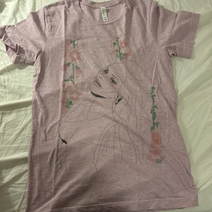 Lana Del Rey rosa t-shirt med tryck - Rosa t-shirt från Canvas med tryck inspirerat av Lana Del Reys album 'Tunnel under Ocean Blvd'. Framsidan har en konstnärlig linjeteckning med blommor i grönt och rosa. Baksidan har albumtitel i rosa och svart text. Klassisk passform och rund hals. Köpt från hennes officiella hemsida, använt ett par gånger men säljer pga kommer ej till tillräckligt mycket användning. Bara skrynklig från att ha legat i garderoben ett tag