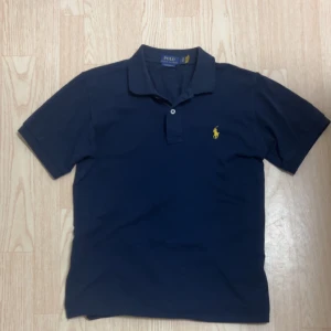  Marinblå Polo Ralph Lauren t-shirt - Snygg marinblå t-shirt från Polo Ralph Lauren i slim fit-modell. Klassisk piké med korta ärmar, krage och två knappar framtill. Ikoniska gula broderade loggan på bröstet. Tillverkad i bomull för en skön känsla.
