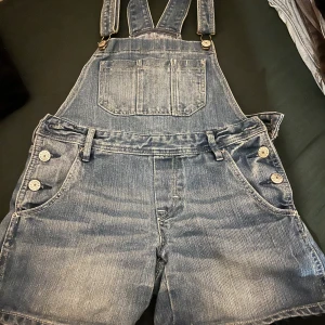 Jeansshorts hängselshorts från H&M - Snygga blå jeansshorts med hängslen från H&M. Klassisk denimlook med fickor fram och bak, dekorativa knappar i sidorna och bröstficka. Perfekt för en chill och avslappnad stil under sommaren.